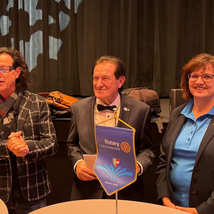 Drei Menschen stehen lächelnd in der Nähe eines Tisches. Der Mann in der Mitte hält ein Banner mit der Aufschrift 'Rotary Club Furstenfeld'. Sie befinden sich in einem Raum mit einem dunklen Vorhang im Hintergrund.