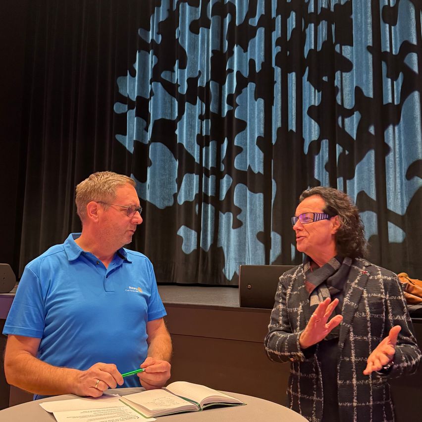 Ein Mann und eine Frau stehen sich gegenüber an einem Tisch, mit einem offenen Buch vor ihnen. Der Mann trägt ein blaues Hemd und eine Brille, während die Frau eine karierte Jacke und eine Brille trägt. Sie stehen vor einem blauen Vorhang mit einem baumartigen Muster.