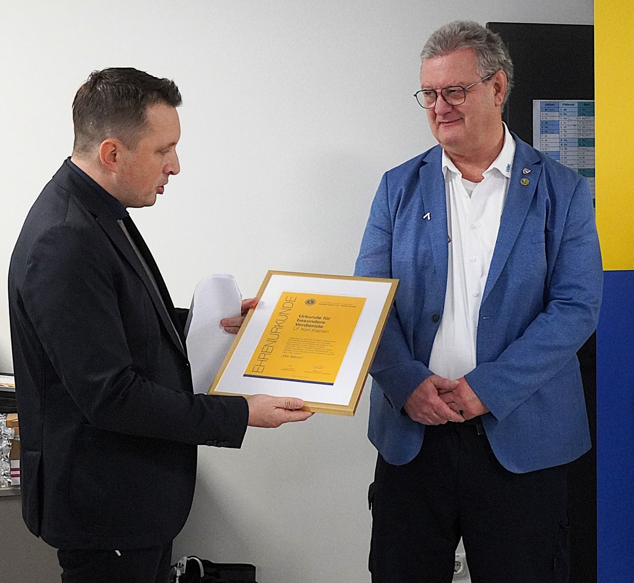 Ein Mann in einem schwarzen Anzug erhält von einem Mann in einem blauen Anzug ein Zertifikat, neben einer Tafel stehend.