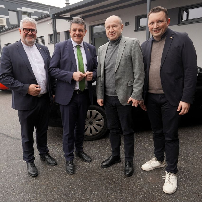 Vier Männer in formeller Kleidung, einschließlich Anzüge und Krawatten, stehen vor einem Auto. Sie scheinen für ein Foto zu posieren. Dahinter befindet sich ein Gebäude mit Glasfenstern.