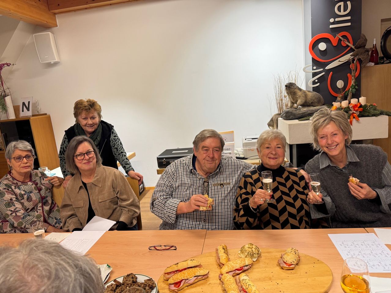 Eine Gruppe von Menschen versammelt sich um einen Tisch mit Sandwiches und Getränken. Sie scheinen eine Mahlzeit gemeinsam zu genießen. Der Tisch ist mit Sandwiches beladen, und eine Frau hält ein Glas Champagner. Ein Mann in der Mitte isst ein Sandwich. Eine Wand im Hintergrund zeigt ein Murmeltier und ein Logo.