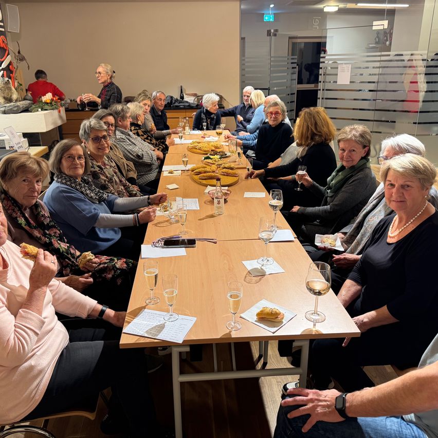 Eine Gruppe von Menschen sitzt um einen langen Tisch herum und genießt Essen und Getränke. Gläser Wein und Snacks stehen auf dem Tisch. Der Raum hat ein modernes Design mit Glaswänden und einer weißen Decke.