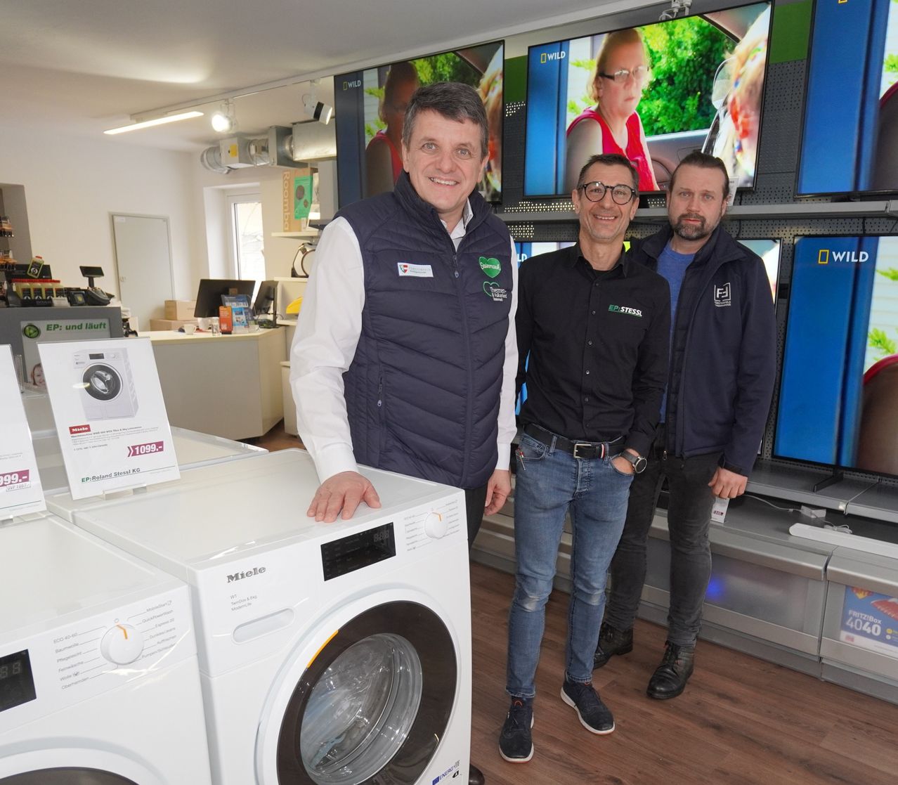 Drei Männer stehen in einem Geschäft in der Nähe von Waschmaschinen, zwei lächeln und einer schaut in die Kamera. Dahinter befinden sich Regale mit Monitoren und einer Person auf dem Bildschirm.