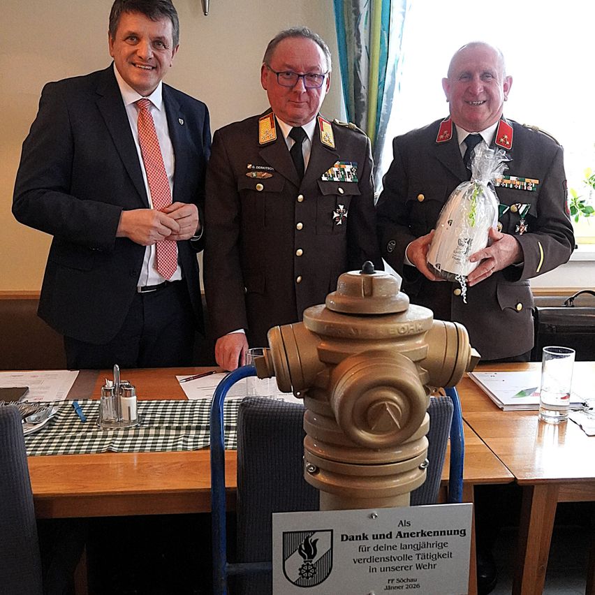 Drei Männer in Uniformen stehen hinter einem Tisch. Der Mann in der Mitte hält ein verpacktes Geschenk. Ein anderer Mann hält einen Feuerhydranten und der dritte Mann steht neben ihnen.