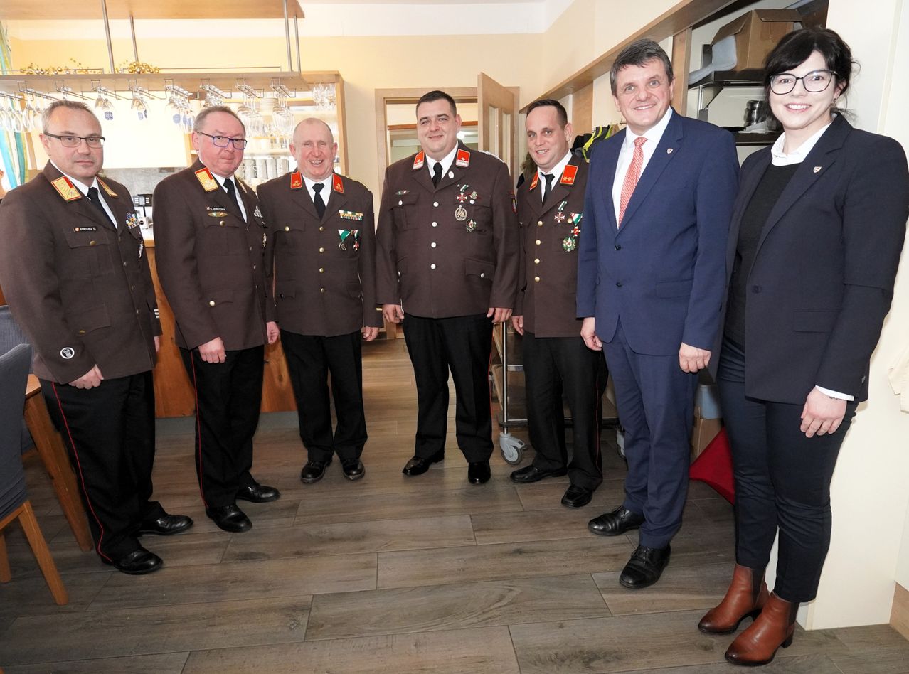 Eine Gruppe von Männern und einer Frau in formeller Kleidung stehen in einem Raum, wahrscheinlich einer Besprechung. Sie lächeln und scheinen für ein Foto zu posieren.