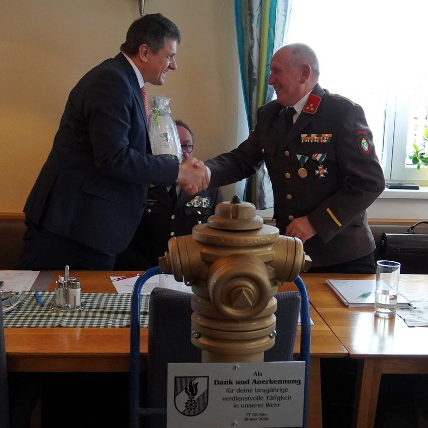 Drei Männer in Anzügen und Uniformen schütteln sich in einem Besprechungsraum die Hände. Ein Schild steht 'Dank und Anerkennung'. Ein Feuerwehrhydrant steht auf dem Tisch. Papiere und ein Glas Wasser sind auf dem Tisch.