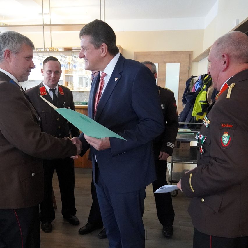 Mehrere Männer in Uniformen, einer schüttelt einem Mann in Anzug die Hand, in einem Raum mit Holzboden und Regalen mit Flaschen.