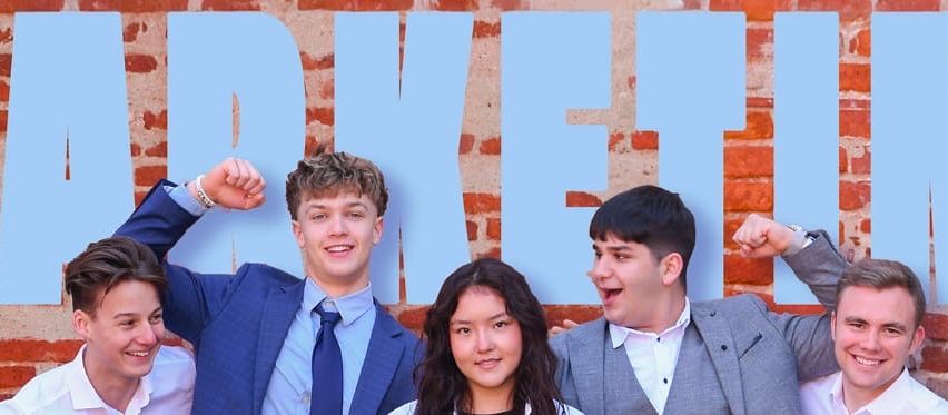 Five young adults are posing for a photo in front of a brick wall with large blue letters spelling out 'MARKET'. They are all smiling and some have their arms raised.