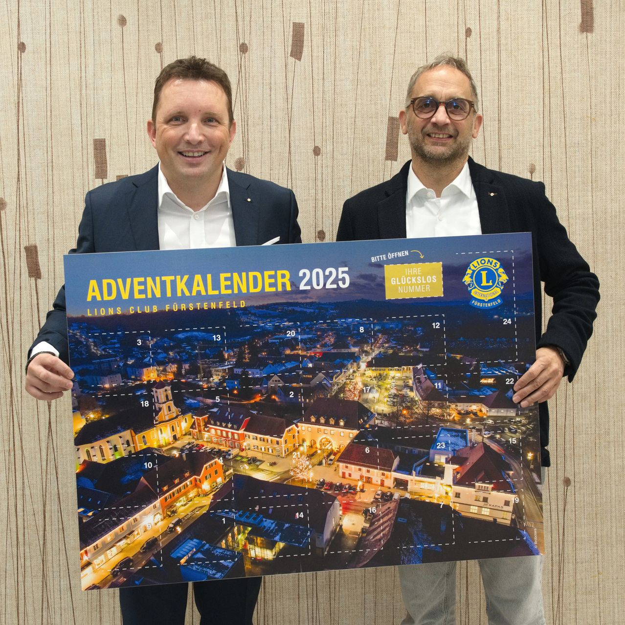 Two men hold up a 2025 Lions Club Fürstenfeld advent calendar, featuring a nighttime cityscape. The calendar has 24 windows, each numbered and marked with a date.