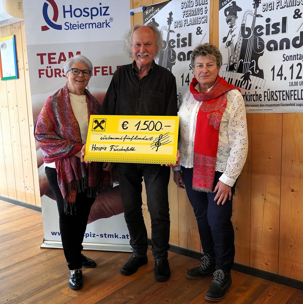 Three individuals stand together, holding a large yellow check for €1500. They are all smiling. Behind them is a wall with a poster advertising a musical event.