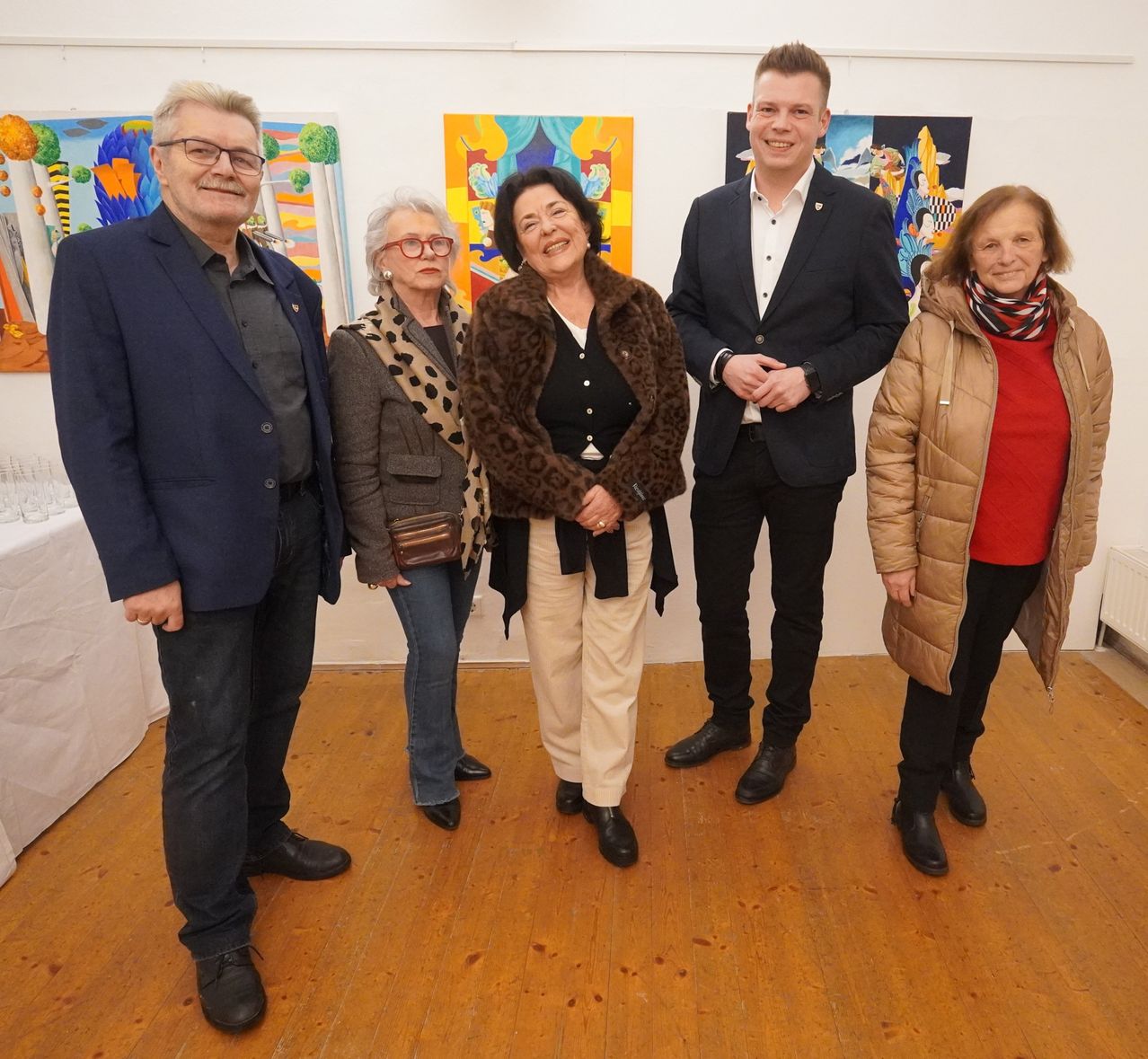Five people stand together in an art gallery, smiling and posing for a photo. Behind them, several paintings hang on the wall.