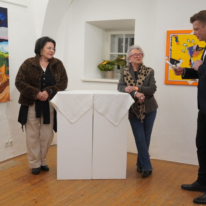 Three people stand in a room with art. Two women are standing near a table, while a man holds papers. The women wear coats and glasses.