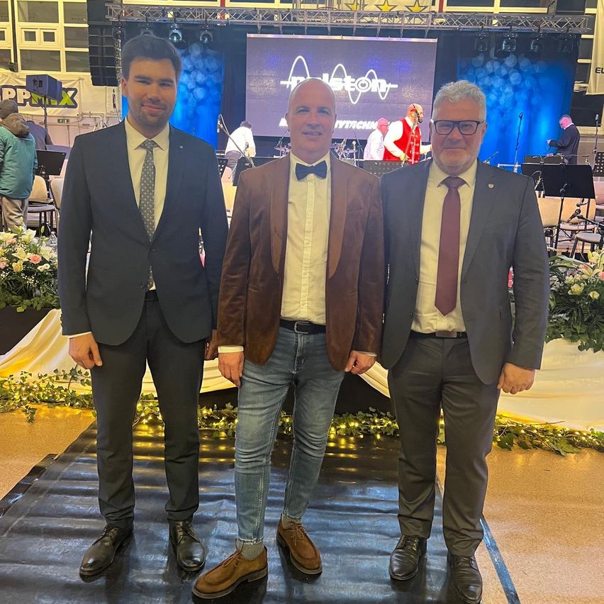 Three men dressed in formal attire stand on a stage, likely posing for a photo. Behind them is a stage with a screen and a musical band.