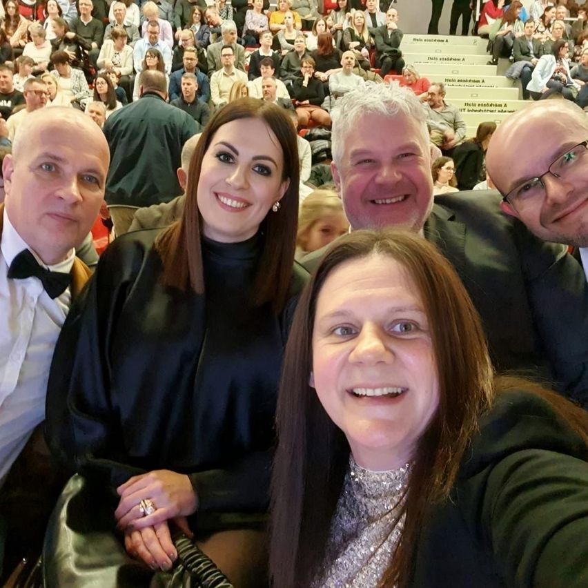Five people are smiling at the camera in an auditorium, two men and three women, with a crowd in the background.