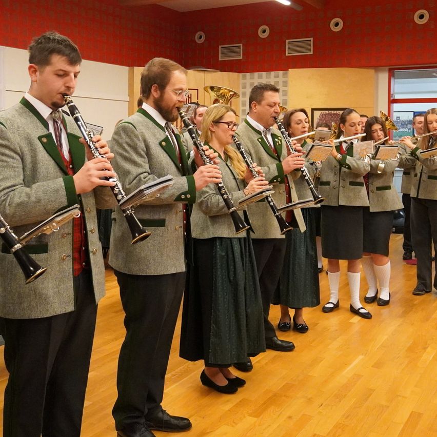 A group of musicians stands in a row on a wooden floor, playing various wind instruments. They wear uniforms with green trim and are accompanied by a few onlookers. The room has red walls and an open window.