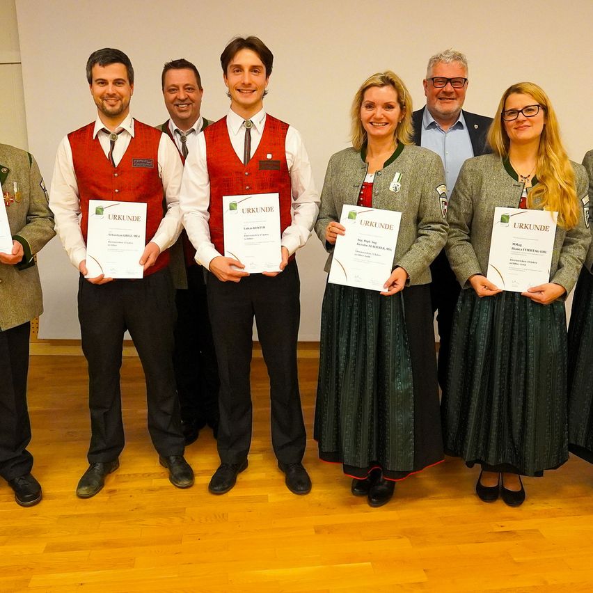 A group of people in traditional attire stand together, smiling and holding certificates. They include two women and three men. Behind them, a man in a suit and glasses completes the ensemble.