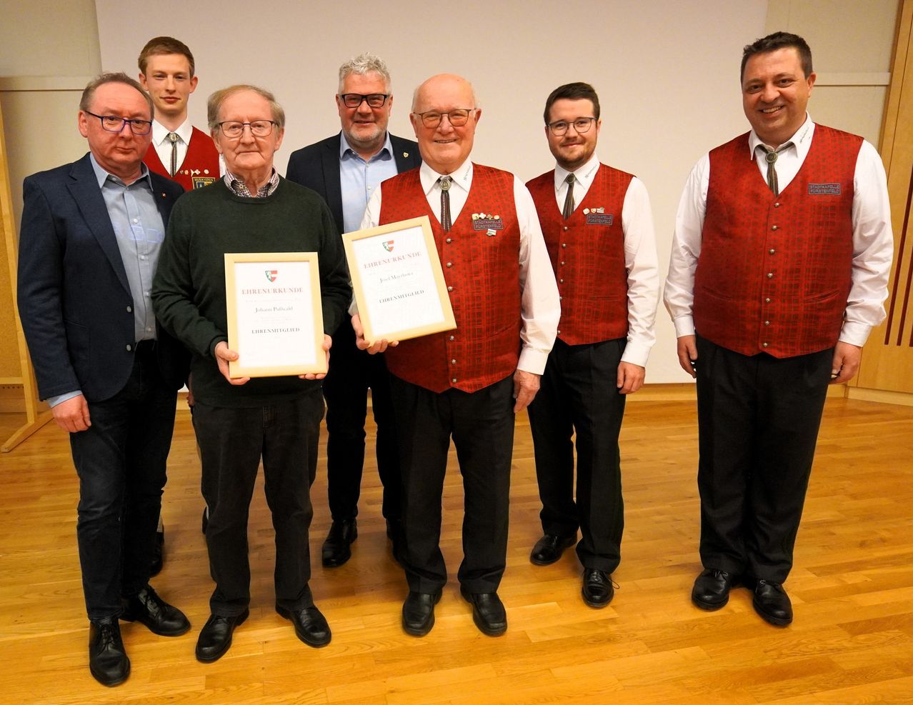 A group of men are standing in a row. They are dressed in formal attire, with two men holding certificates. The man on the right is smiling and wearing a red vest.