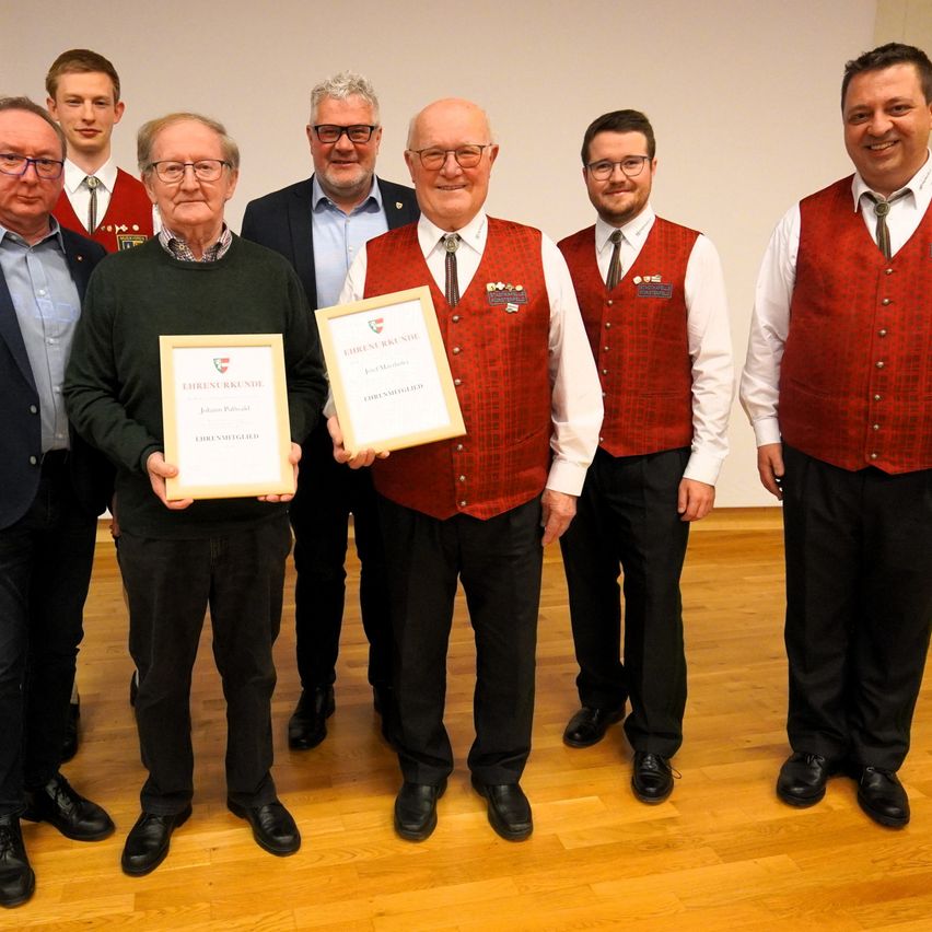 Seven men in formal attire stand on a wooden floor. Two men in front hold certificates, while others wear red vests with emblems. They all wear glasses and ties. The background is white.