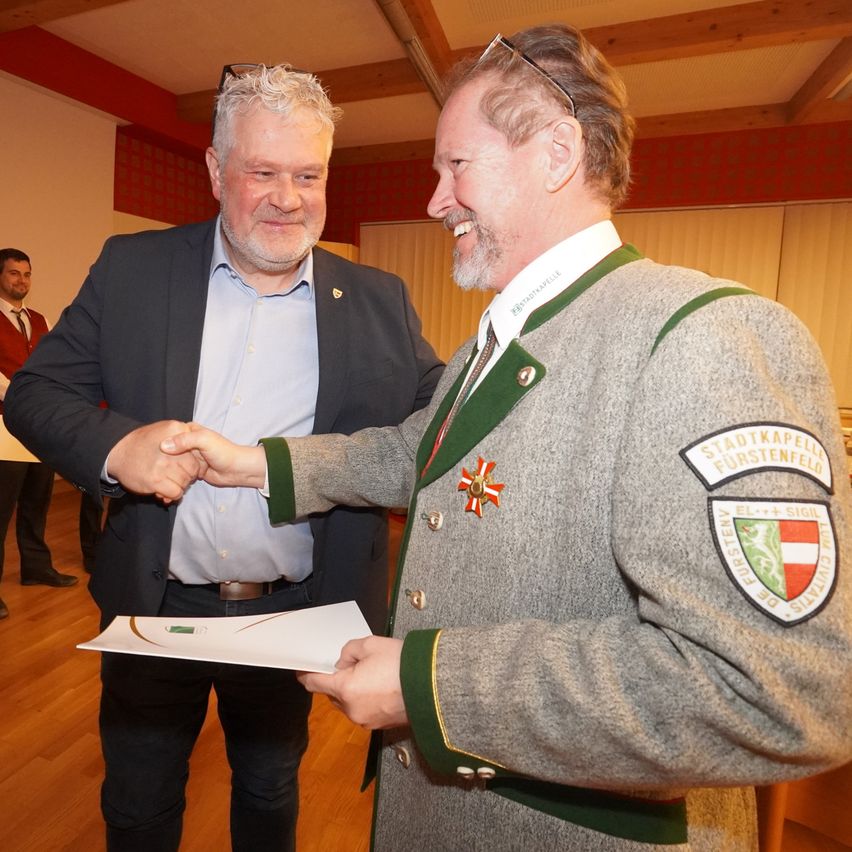 Two men are shaking hands in a room, one wearing a traditional German uniform. A paper is being handed to the uniformed man. A man in a suit stands behind them.