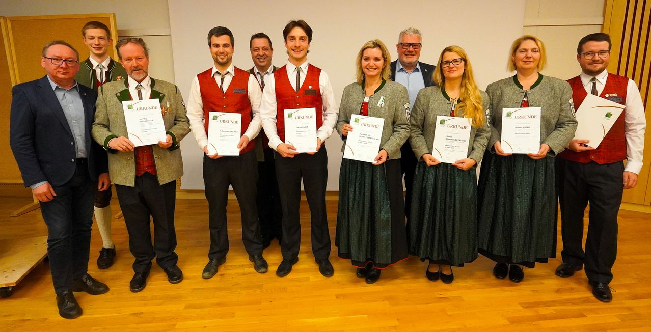 A group of people, both men and women, dressed in traditional attire, stand together holding certificates. They are posing for a photo in a formal setting.