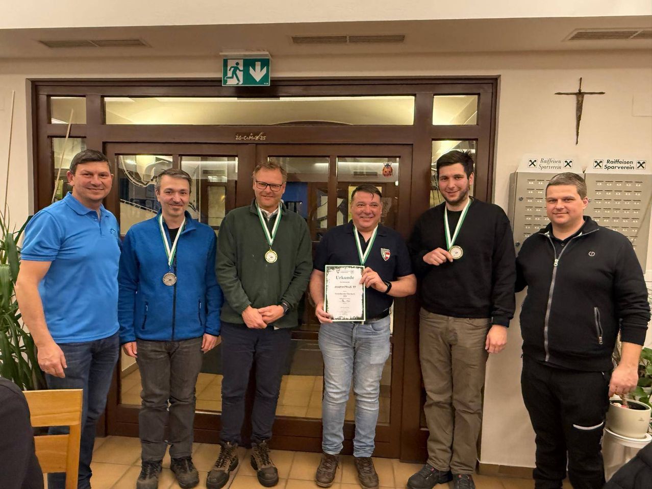Five men stand in front of a door, smiling for a photo. The man in the center holds a certificate and a medal. The others wear medals as well. Behind them, an emergency exit sign is mounted on the wall.