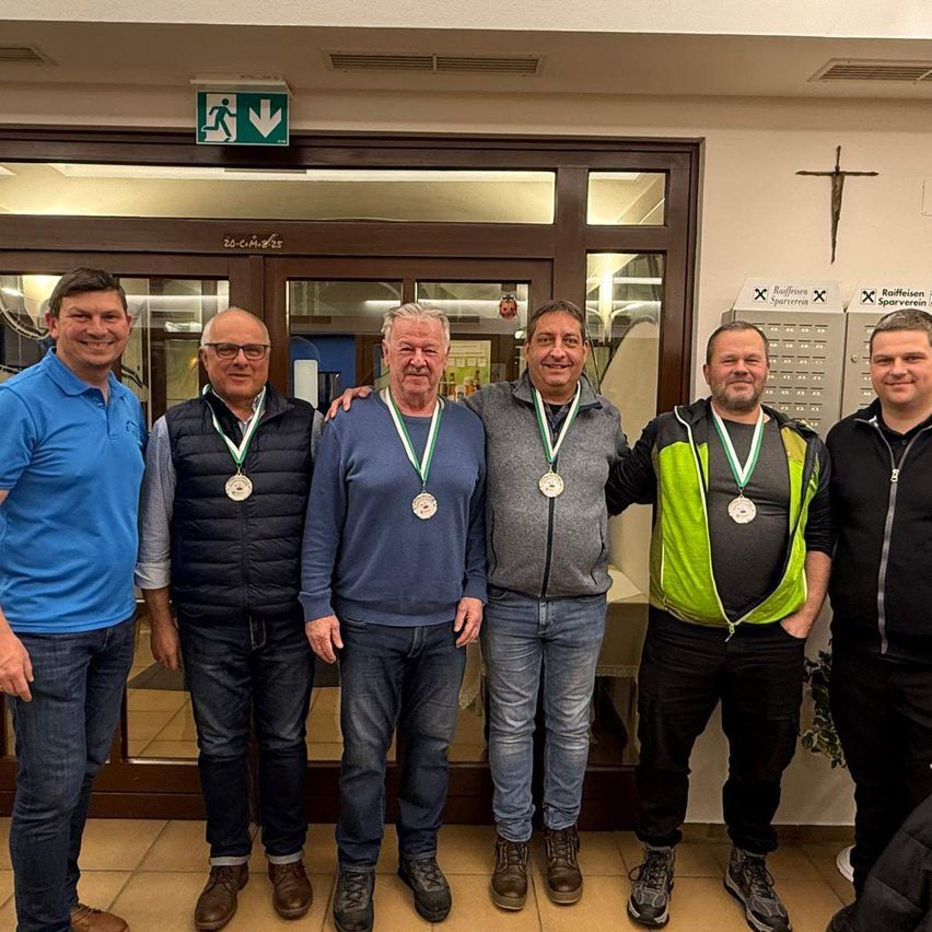 Five men wearing medals stand together in front of a building. They are smiling and appear to be posing for a photo. The man on the left wears a blue polo shirt, and the man on the right wears a black jacket.