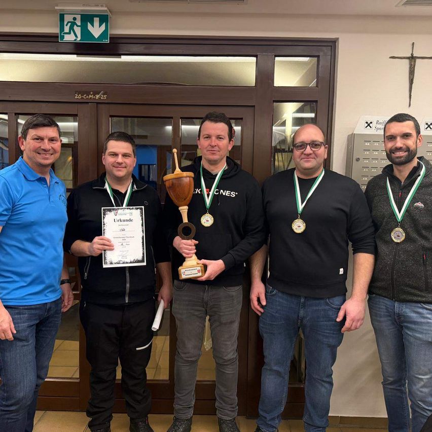 Five men stand in front of a door. The man on the left wears a blue shirt, and the man in the middle holds a trophy and certificate. They all wear medals. Behind them is a door with a cross symbol and an exit sign.