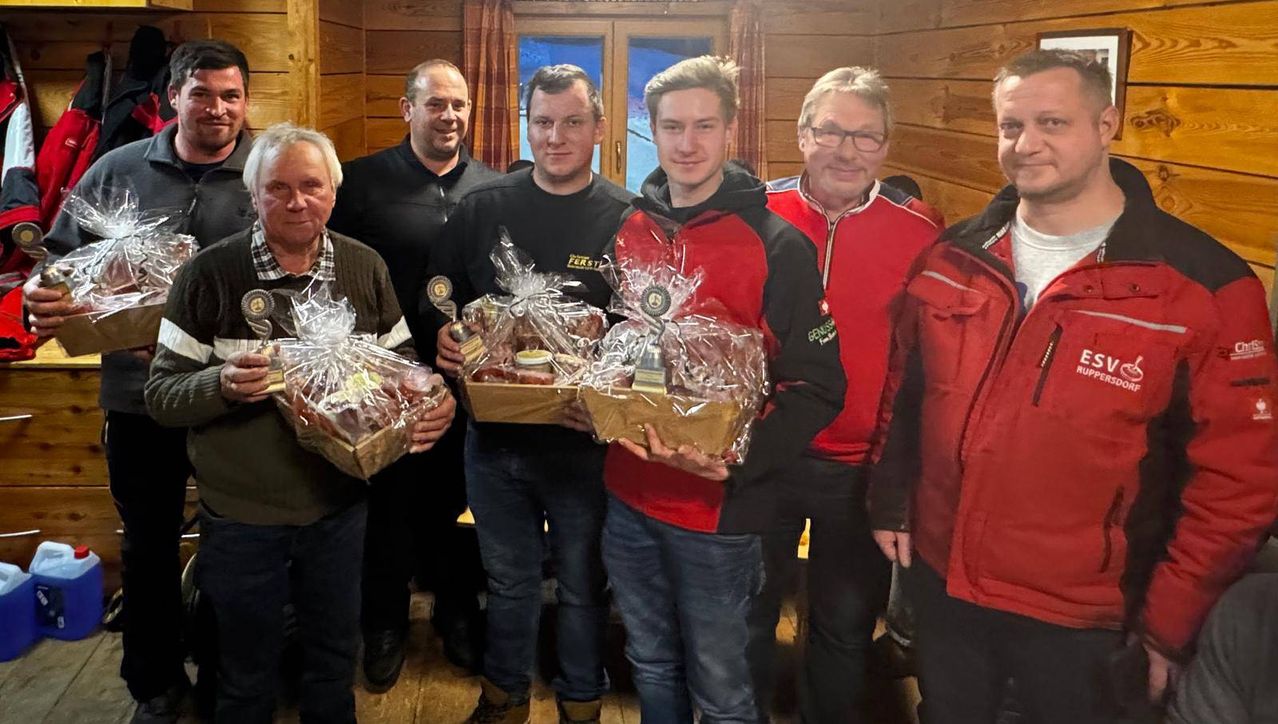 Five men stand in a room with wooden walls, holding gift baskets. They all smile and appear to be posing for a photo. The man in the middle holds a trophy.