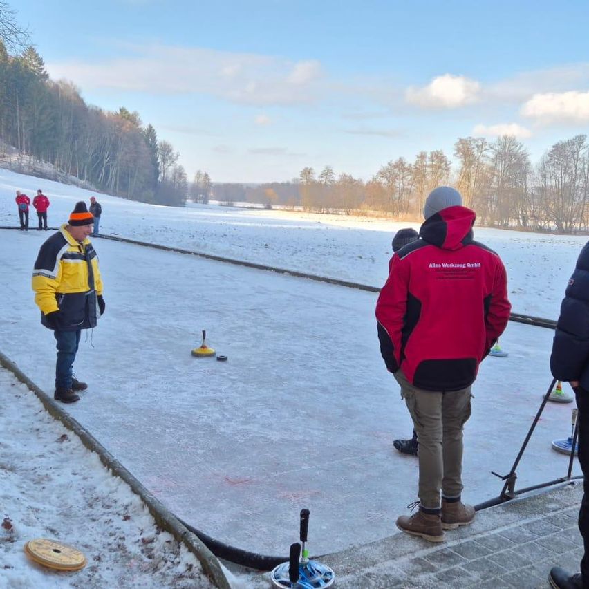 A group of people are on an ice rink, one wearing a red jacket and hat, likely playing a game. The ice rink is surrounded by snow and trees.