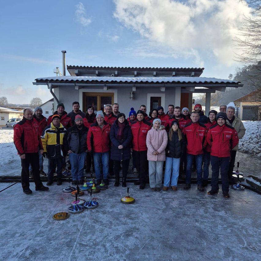 A group of people in red jackets pose for a photo on an ice rink with a house in the background.