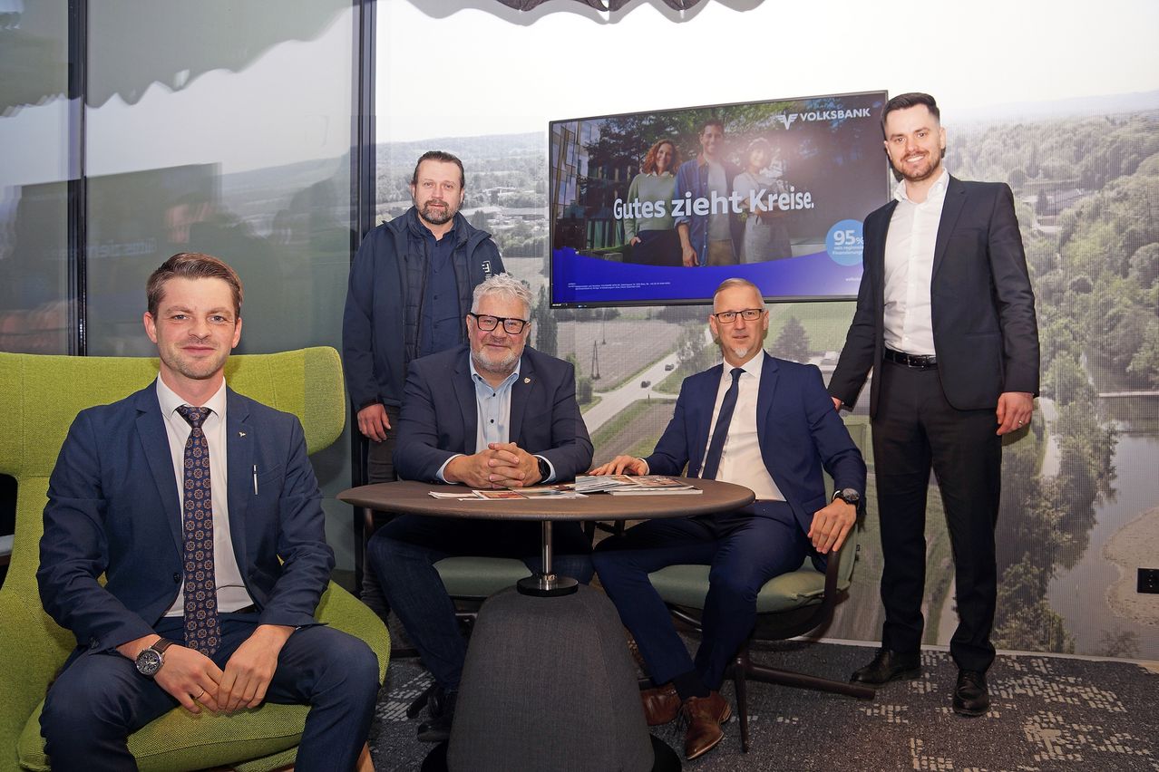 Fünf Männer in Anzügen sitzen in einem Büro mit einer großen Volksbank-Werbung an der Wand hinter ihnen. Sie scheinen in ein Gespräch vertieft zu sein.