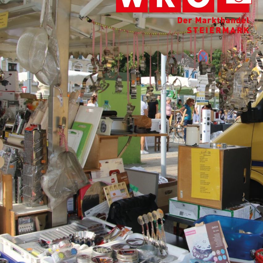 A market stall displays various items for sale. The stall is organized with items such as a yellow box, a box of cookies, and a box of tea bags. There are also kitchen utensils, plastic containers, and a box of cookies. There are people walking around the market.