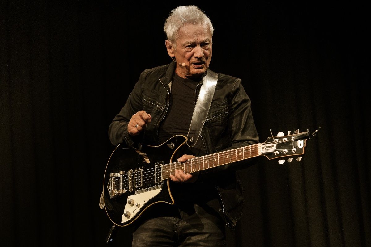 Ein älterer Mann steht auf der Bühne und spielt eine schwarze E-Gitarre. Er trägt einen schwarzen Mantel und hat ein verkabeltes Mikrofon.