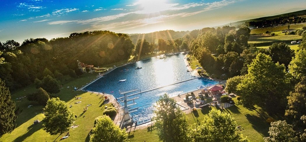 Ein Luftbild eines Freibades, umgeben von üppiger Grünfläche, Bäumen und Bergen. Mehrere Menschen sonnen sich und schwimmen, während andere unter Sonnenschirmen sitzen.