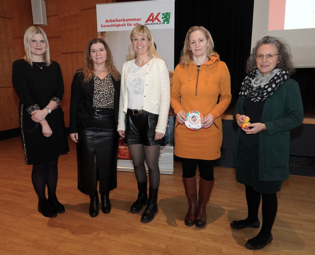 Fünf Frauen stehen lächelnd für ein Foto zusammen. Die Frau in der Mitte hält ein farbenfrohes Windrad. Hinter ihnen ist ein weißes Banner mit dem Text Arbeiterkammer Gerechtigkeit fur alle.