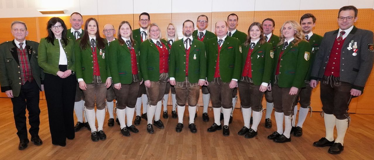 Eine Gruppe von Menschen in traditioneller bayerischer Kleidung posiert für ein Foto. Sie tragen grüne Jacken und braune Hosen. Die Frauen tragen rote Westen mit weißen Schleifen, während die Männer schwarze Krawatten tragen.