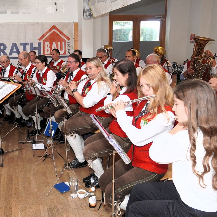 Eine Blaskapelle spielt in einem Raum. Einige von ihnen spielen Flöte, andere Trompete. Sie tragen alle rote Westen.