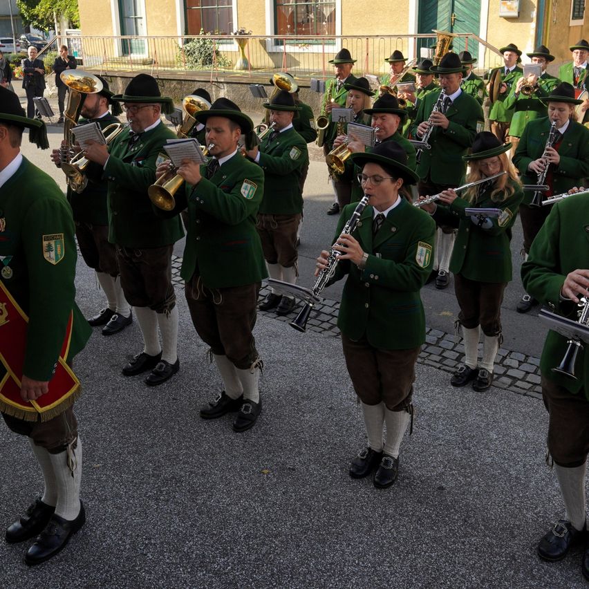 Eine Blaskapelle in grünen Uniformen spielt verschiedene Instrumente, darunter Trompeten, Posaunen und Klarinetten, bei einer Straßenaufführung vor einem Gebäude.