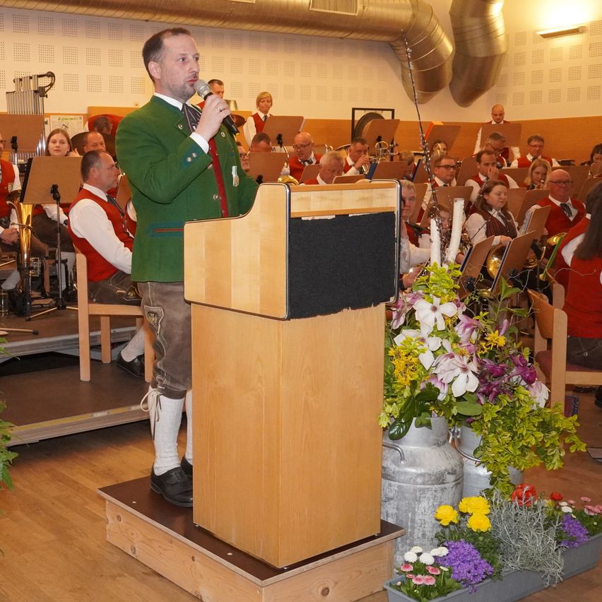 Ein Mann in einer grünen Jacke und weißen Socken steht an einem Podium und spricht in ein Mikrofon. Hinter ihm spielt ein Orchester mit verschiedenen Instrumenten. Der Raum ist ein Saal im Inneren, mit Blumen und Holzkisten auf dem Boden.