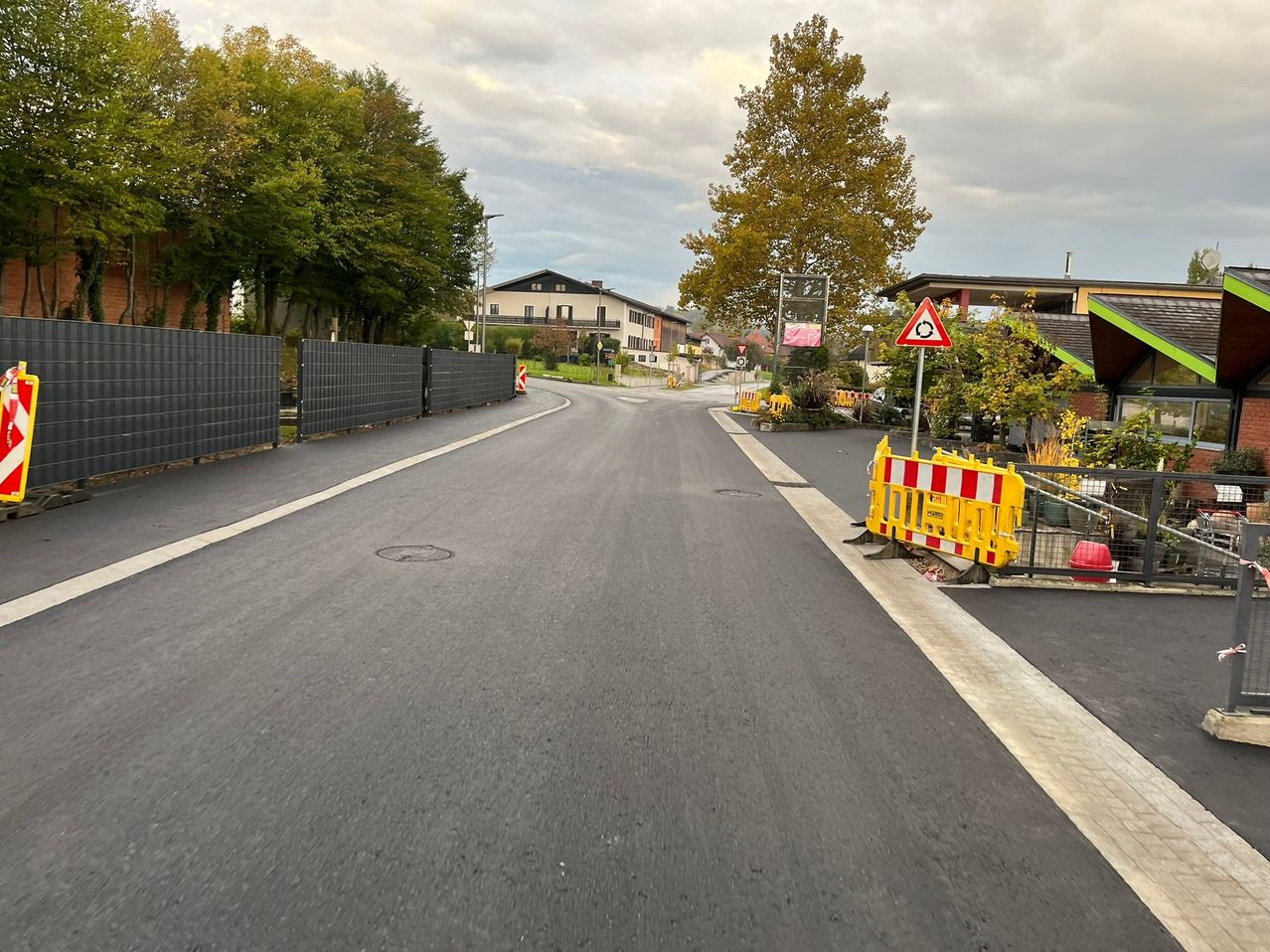 Eine leere Straße mit neuem Asphalt, gesäumt von Zäunen und Bäumen, mit einem Verkehrsschild und einer Absperrung auf der rechten Seite. Gebäude und Straßenlaternen sind im Hintergrund zu sehen.