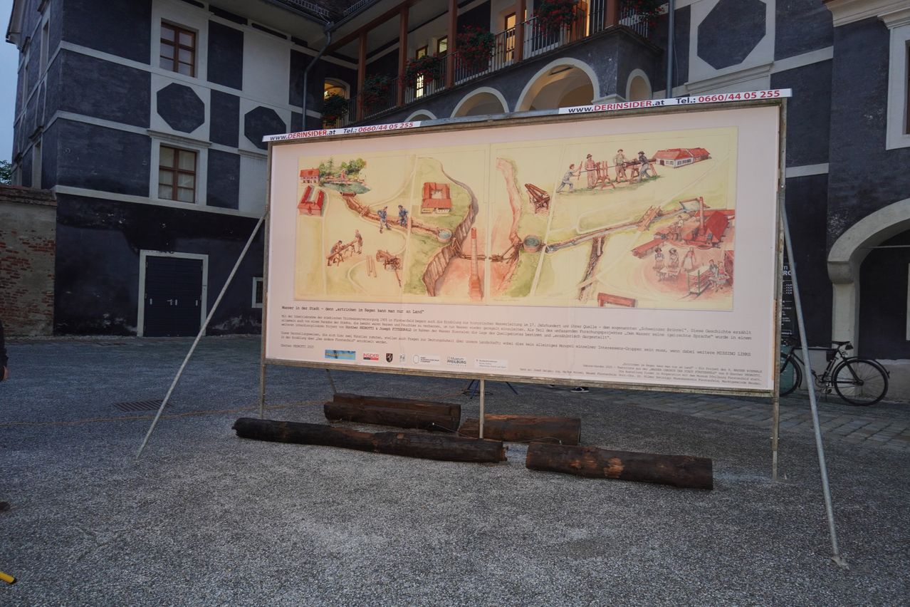 Ein großes Plakat, das einen historischen Stadtplan zeigt, ist im Freien vor einem Gebäude ausgestellt. Das Plakat hat eine Karte der Stadt mit Menschen und Gebäuden. Darunter sind mehrere Holzstämme auf dem Boden platziert.