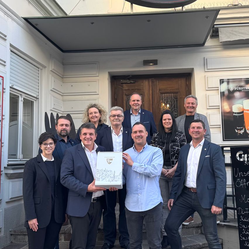 Eine Gruppe von Menschen steht lächelnd vor einem Gebäude. Zwei Männer halten ein gerahmtes Zertifikat. Dahinter sind eine Tür und ein Schild mit einem Bierbild zu sehen.