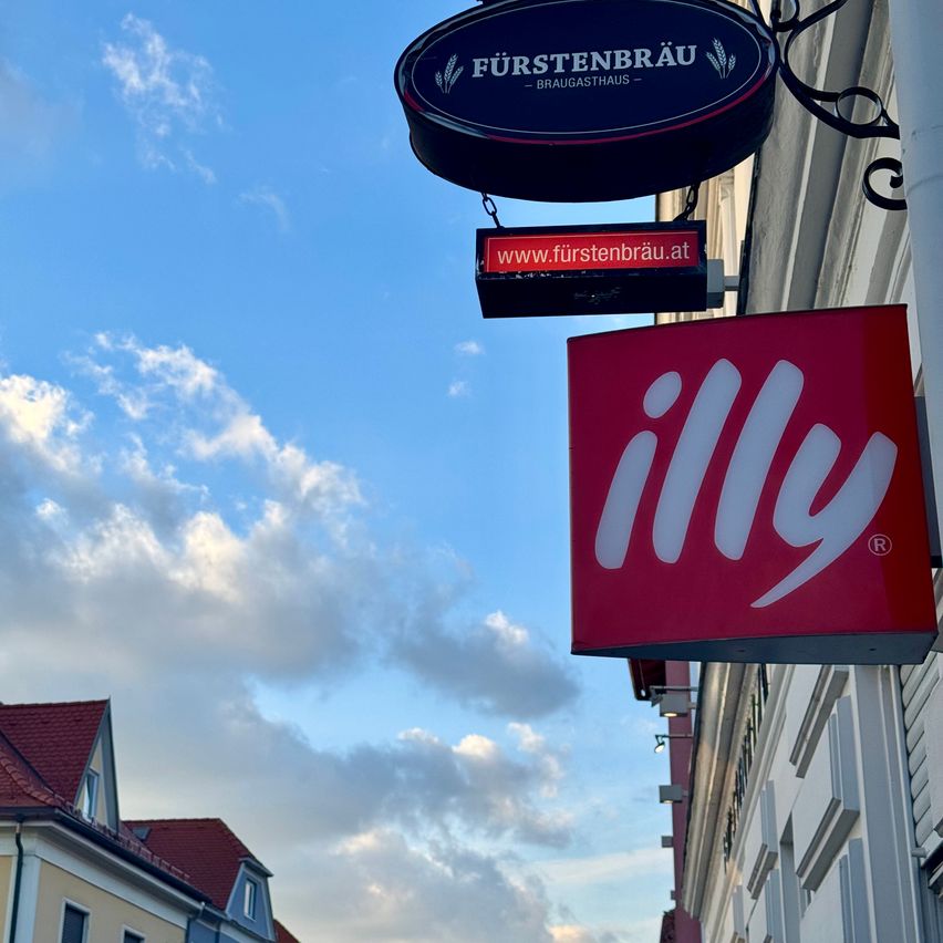 Ein rotes Schild für illy Kaffee hängt vor einem Gebäude mit einem blauen Fürstenbräu-Schild darüber, vor einem klaren blauen Himmel mit verstreuten Wolken.