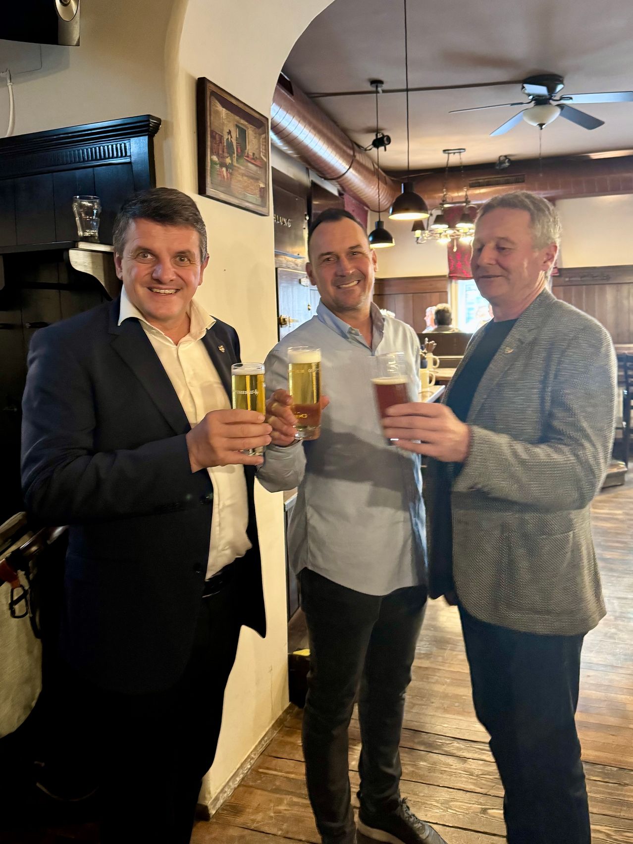 Drei Männer stehen zusammen in einer Bar, halten Biergläser hoch und lächeln. Der Mann auf der linken Seite trägt einen dunklen Anzug, der Mann in der Mitte ein hellblaues Hemd und der Mann auf der rechten Seite einen grauen Blazer.