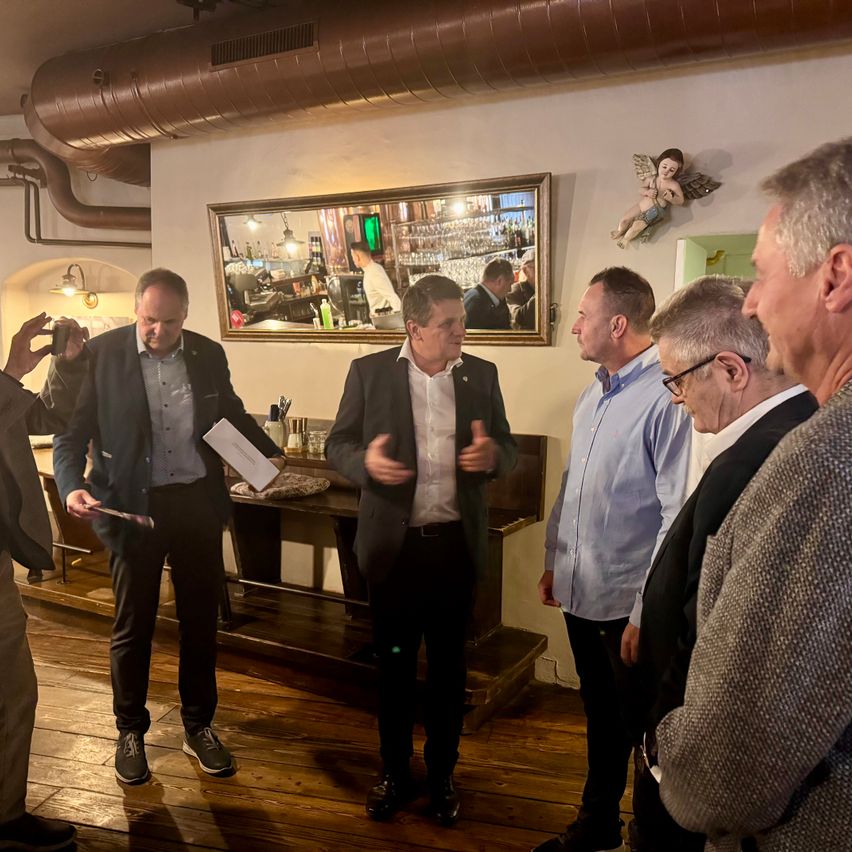 Mehrere Männer in formeller Kleidung befinden sich in einem Raum mit einer Bar. Ein Mann gestikuliert und spricht, während andere zuhören. Ein Spiegel reflektiert eine Bar mit Menschen darin.