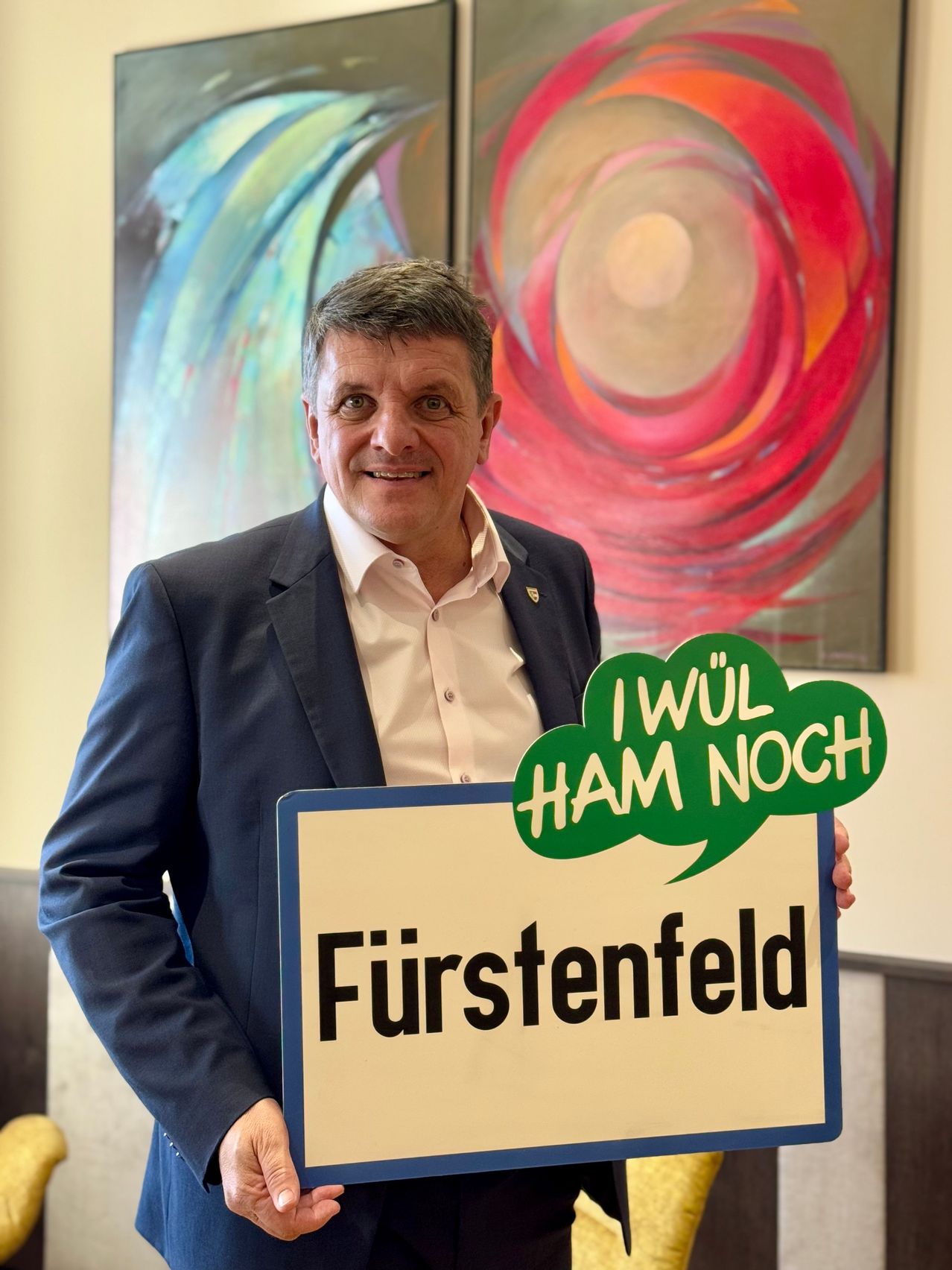 Ein Mann steht in einem Raum und hält ein Schild mit der Aufschrift 'Furstenfeld' und einer grünen Sprechblase, die 'I WÜL HAM NOCH' sagt. Er lächelt und trägt einen Anzug. Hinter ihm ist ein abstraktes Gemälde.