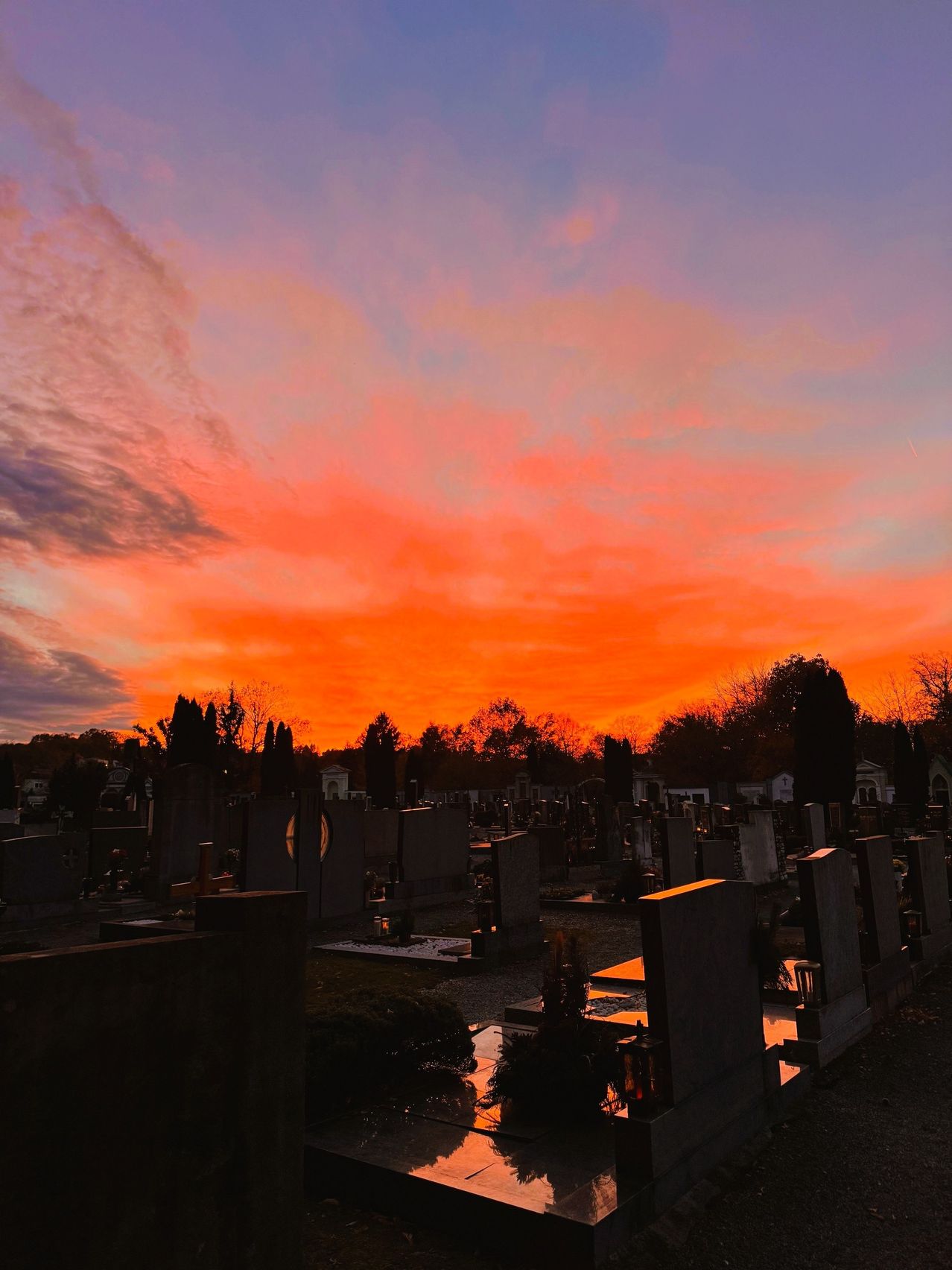 Ein Friedhof bei Sonnenuntergang mit leuchtenden orangen und rosa Farbtönen am Himmel, umgeben von Bäumen und Grabsteinen.