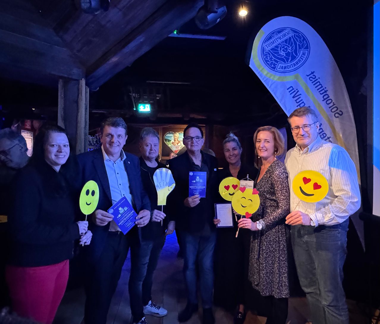 Eine Gruppe von Menschen steht lächelnd für ein Foto vor einem Banner, das hinter ihnen hängt und hält Smiley-Gesicht-Requisiten. Sie sind in einem dunklen Raum mit einem Logo und Text auf dem Banner.