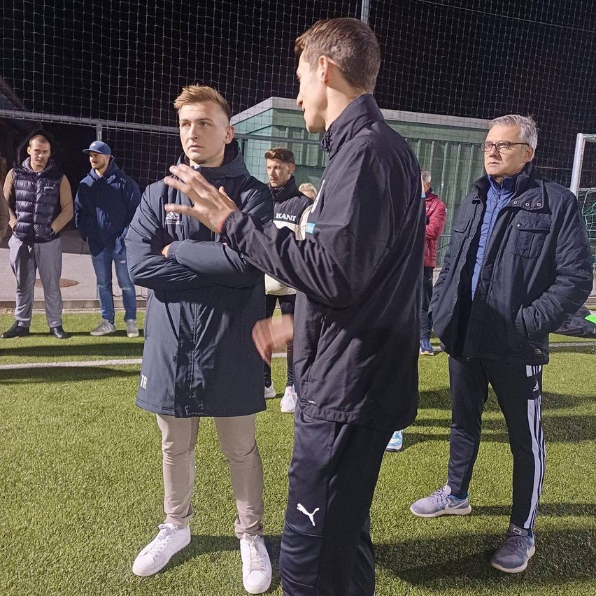Eine Gruppe von Männern steht auf einem Fußballfeld und führt ein Gespräch. Der Mann in der Mitte trägt eine schwarze Jacke mit dem Wort 'Puma' darauf. Hinter ihm lauschen ein Mann mit Brille und ein weiterer Mann aufmerksam. Im Hintergrund stehen weitere Männer und beobachten das Gespräch.