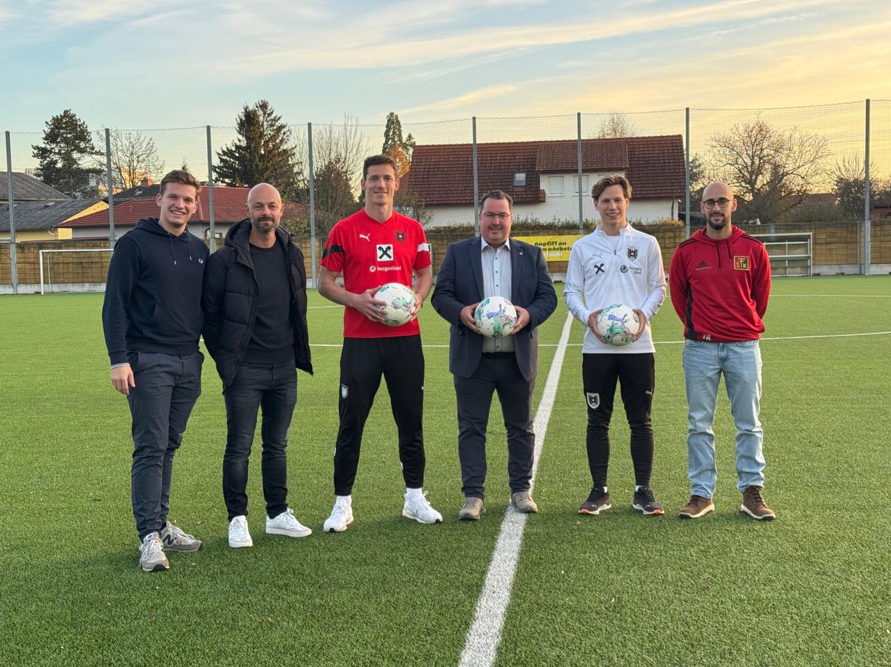 Fünf Männer stehen auf einem Fußballfeld mit einem Banner dahinter. Drei halten Fußballbälle.